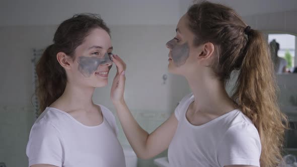 Side View Smiling Young Woman Applying Face Mask on Twin Sister Talking alt