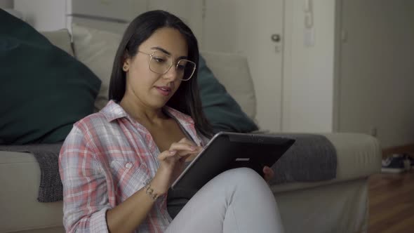Concentrated Young Woman in Eyeglasses Sitting on Floor and Using Tablet. alt