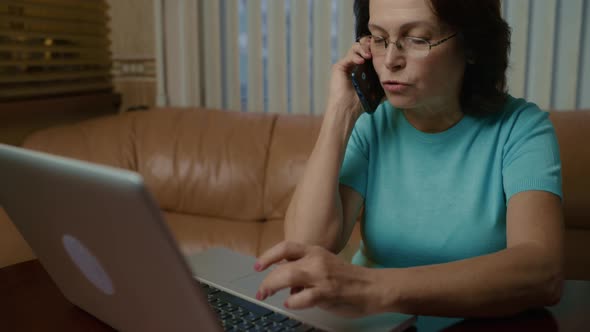 Elderly Woman with Glasses Works on a Laptop Cheerfully Speaks on a Smartphone