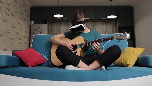Young Man Sitting on Sofa with the Virtual Reality Glasses Is Playing at the Guitar alt