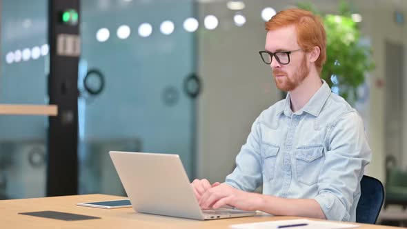 Focused Young Redhead Man Working on Laptop in Office alt