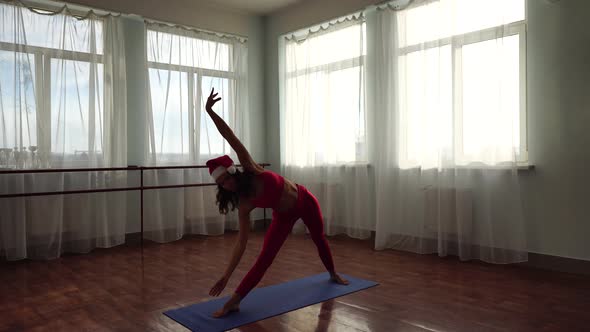 A Young Woman Fitness Instructor in Santas Hat and Red Sportswear Leggings and Top Stretching in the alt