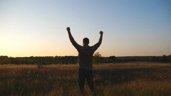 Happy Guy Going Through Grass Field and Victoriously Raising Hands Up at Sunset alt