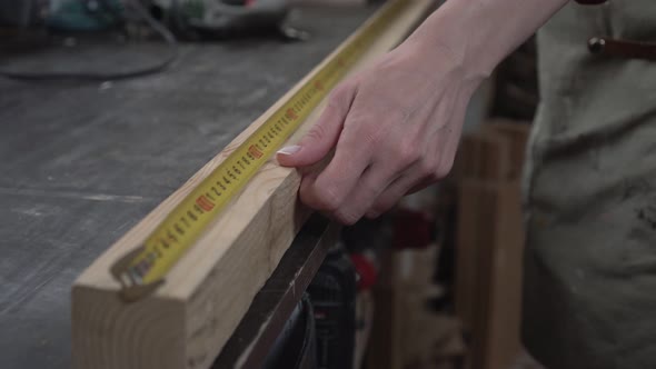 A Woman Carpenter Measures the Length of a Wooden Block on a Workbench with a Tape Measure alt