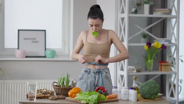 Slim Fit Caucasian Woman Measuring Waist Chewing Salad Leaf Standing at Home Indoors alt