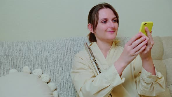 Woman musician with flute looking at mobile phone at home on sofa in living room