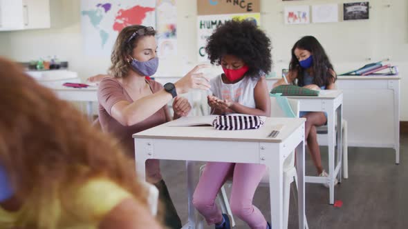 Female teacher and girl wearing face masks sanitizing her hands at school alt
