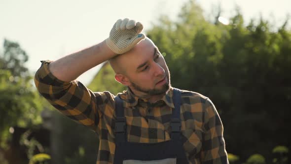 Bearded man in overalls fanning himself with a straw hat, and wiping sweat from his forehead alt