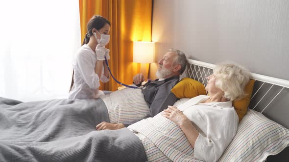Female Nurse Checking Sounds in Heart and Lungs of Sick Senior Woman Lying on Bed
