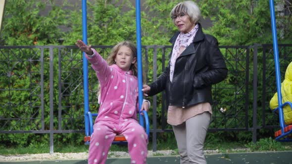 Portrait of Happy Hyperactive Granddaughter Talking with Grandmother Swinging Swings on Playground alt