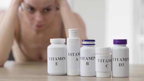Row of Vitamin Bottles on Table with Blurred Caucasian Young Woman at Background Thinking alt