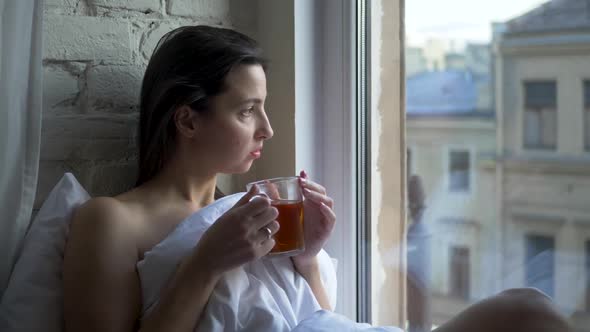 Young Brunette Woman Sits on the Window Wrapped in a White Blanket alt