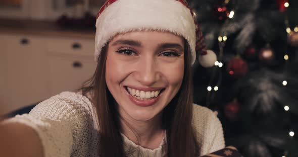 Beautiful Woman Wearing Christamas Hat Sitting Near Christmas Tree at Home Holding and Shaking Gift alt