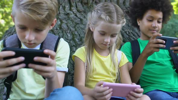 Children Playing Smartphone Games Sitting Under Tree in Park, Gadget Addiction