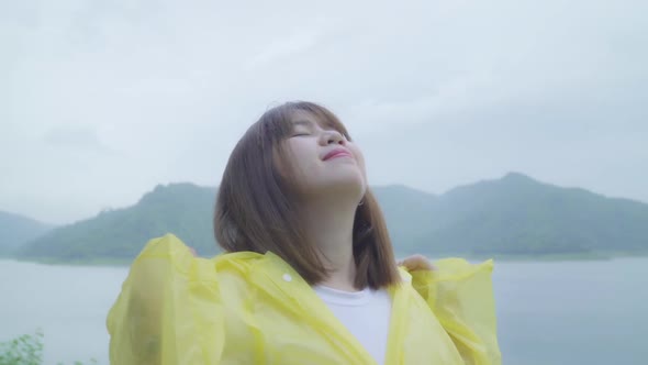 Young Asian woman feeling happy playing rain while wearing raincoat standing near lake. alt