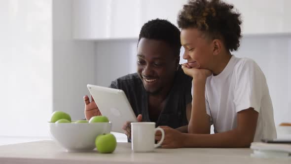 Young Man and Child are Using Tablet and Smiling While Sitting at Table in White Room Spbi alt