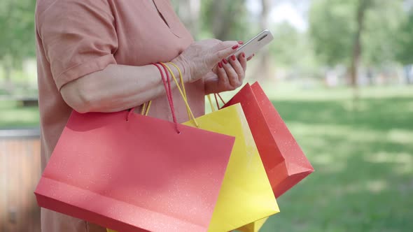 Side View of Unrecognizable Retiree with Shopping Bags Scrolling Social Media on Smartphone. Relaxed alt