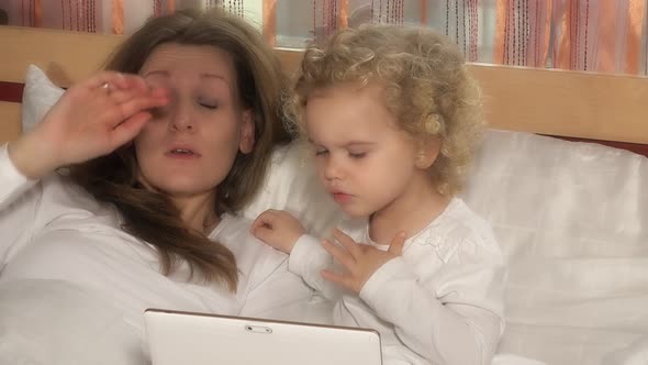 Nice Child Girl Looking at Tablet Computer while Mother Sleeping in Bed
