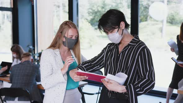 Young Caucasian Female Boss Helps Asian Male Colleague at Work, Both Wearing COVID-19 Masks at alt