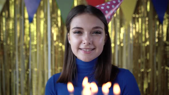 Closeup of a Birthday Cake with Burning Candles in the Hands of a Young Girl in a Birthday Cap alt