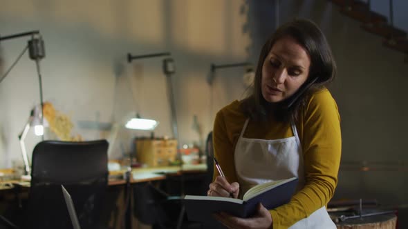 Caucasian female jeweller in workshop wearing apron, sitting at desk, having call and taking order alt