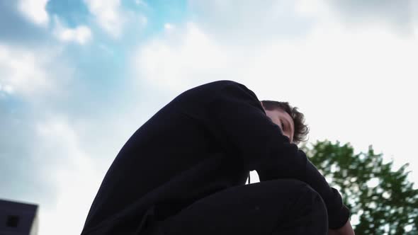Stylish Man in a Black Hoodie Sits in City Park and Looks at Camera alt