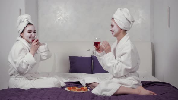 Two Friends Drink Wine and Go Crazy with Glasses Sitting in Robes on the Bed