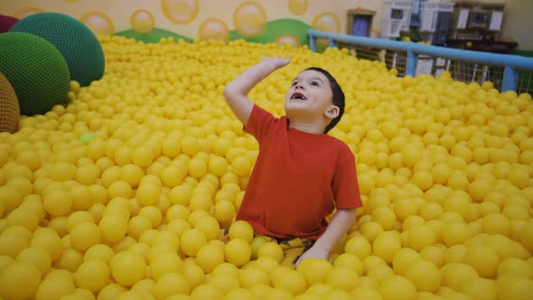 A Little Boy Sits in a Pool with Balls and Throws Balls with His Hand