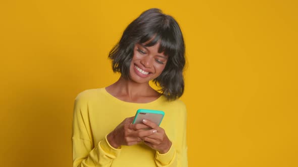 Young Happy Dark Skinned Woman Checks Message on Smartphone Against Orange Wall alt