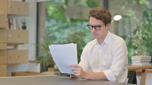 Young Man Reading Documents While Sitting in Modern Office alt