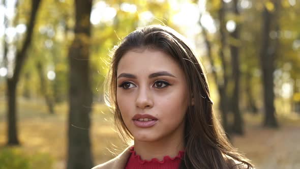 Portrait of Positive Brunette with Makeup Looks at Camera in Sunny Autumn Park alt
