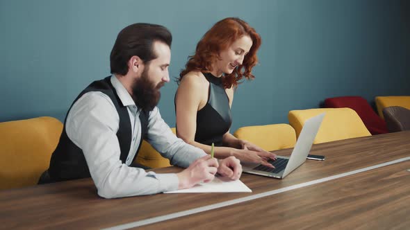 Woman Typing Text on a Laptop in the Office and Next To a Man with a Beard Writing Text alt