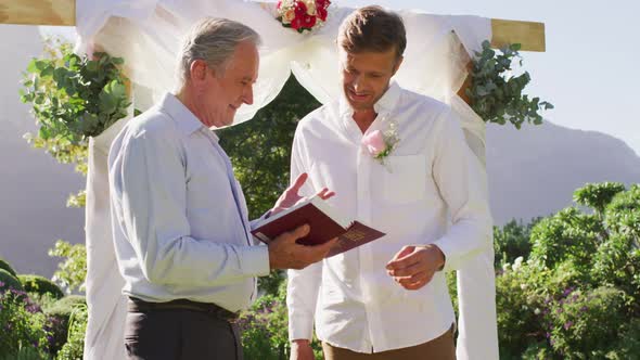 Smiling caucasian senior male wedding officiant holding book and groom standing in outdoor altar alt