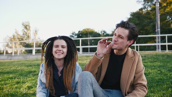 Happy Young Hipster Couple Sitting on Grass and Smoking Marijuana Joint in Park Slow Motion alt
