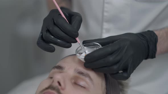 Closeup Beautician Applying White Purifying Enzyme Peel on Face of Young Caucasian Man Lying in alt