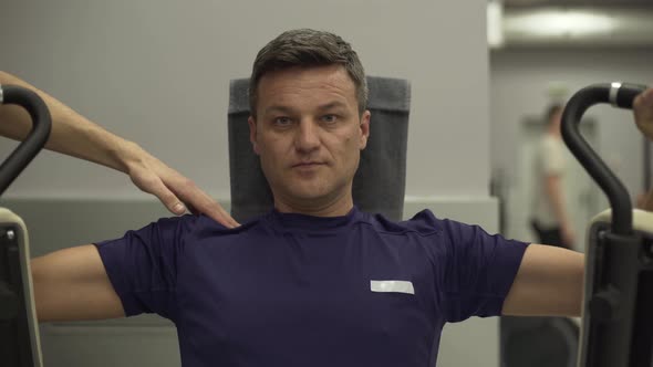 Middle-aged Sportsman Doing Strength Exercises on the Simulator for Hands Looking at Camera alt