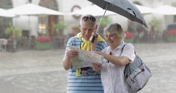 Senior Tourists in Rainy Weather Stand and Looking for the Route in Map alt