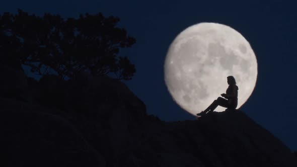 Silhouette of Woman Siting on Rocks Opposite Huge Moon