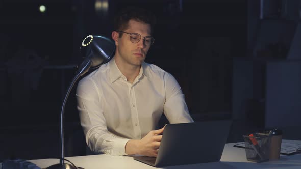 Young Man with Glasses Works at Night at a Computer in a Modern Office
