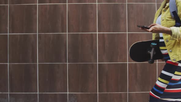 Mixed race woman walking on street holding her skateboard and her phone alt