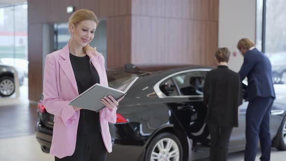 Portrait of Pleasant Woman in Pink Jacket with a Big Book About Cars in Front of Couple Choosing alt