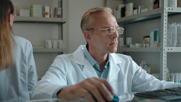 A Mature Pharmacist with Glasses Sitting at a Computer in a Pharmacy alt