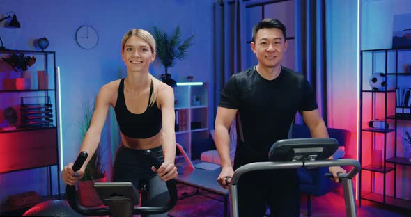 Couple in Sportswears which Exercing on Bike Trainer and Treadmill During Evening Home Workout alt