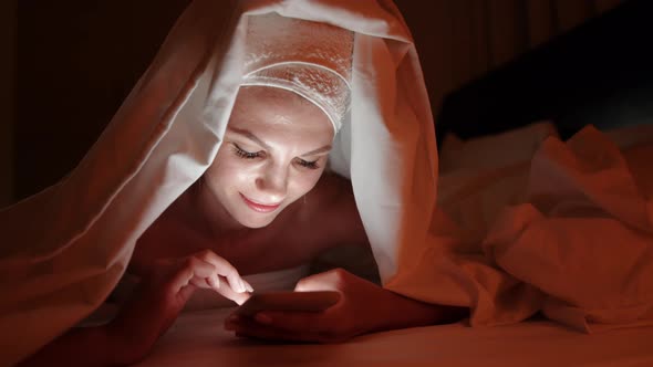 Beautiful Young Woman Using Smart Phone Under Blanket in Bedroom on Bedtime alt