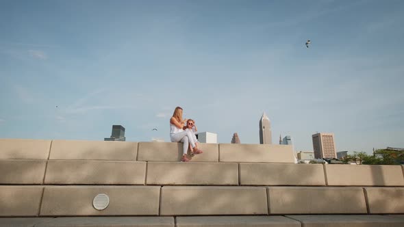 Happy Mother and Daughter are Resting in a City Park on a Bench alt