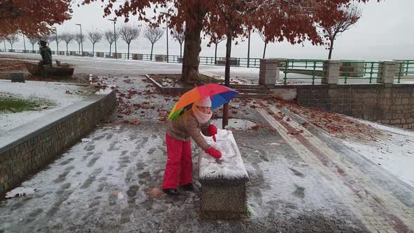 Child girl with colorful umbrella plays outdoor kicking snowball. Slow-motion alt