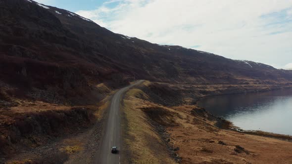 Drone Tracking Car Driving By Fjord And Towards Mountains alt