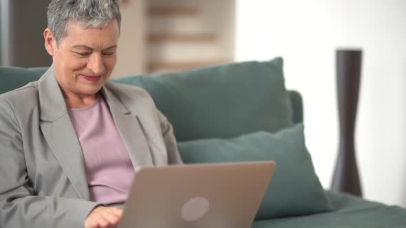 Mature Woman in a Gray Suit and with Short Gray Hair is Having a Video Call Using a Laptop and alt