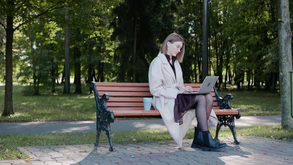 Young Girl Sits on a Park Bench Working on a Laptop and Drinking From an Eco Cup