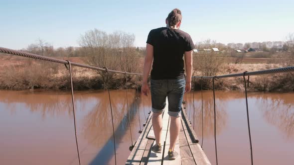 Young Man Is Walking on a Suspended Wooden Bridge Over the River. Back View.